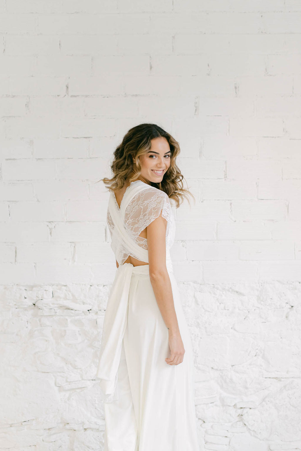 Modelo de espaldas pero mirando y sonriendo a cámara con pelo en movimiento. La modelo lleva el mismo vestido de novia pantalon con straps de encaje blanco en el escote y formado una X en la espalda.