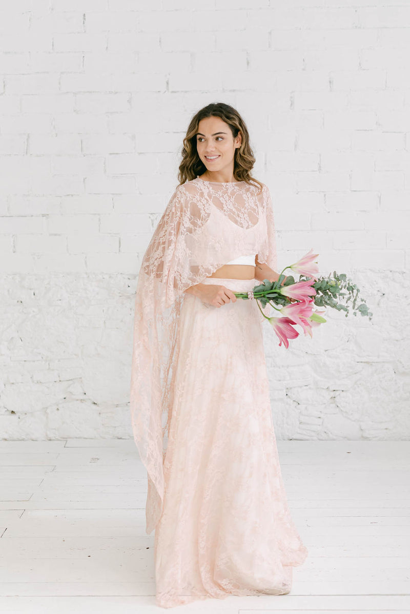 modelo chica mirando hacia la derecha sujetando un ramo de flores rosas en la mano. La modelo lleva un vestido de novia dos piezas con top capa rosalia y falda de encaje de cuarzo rosa.